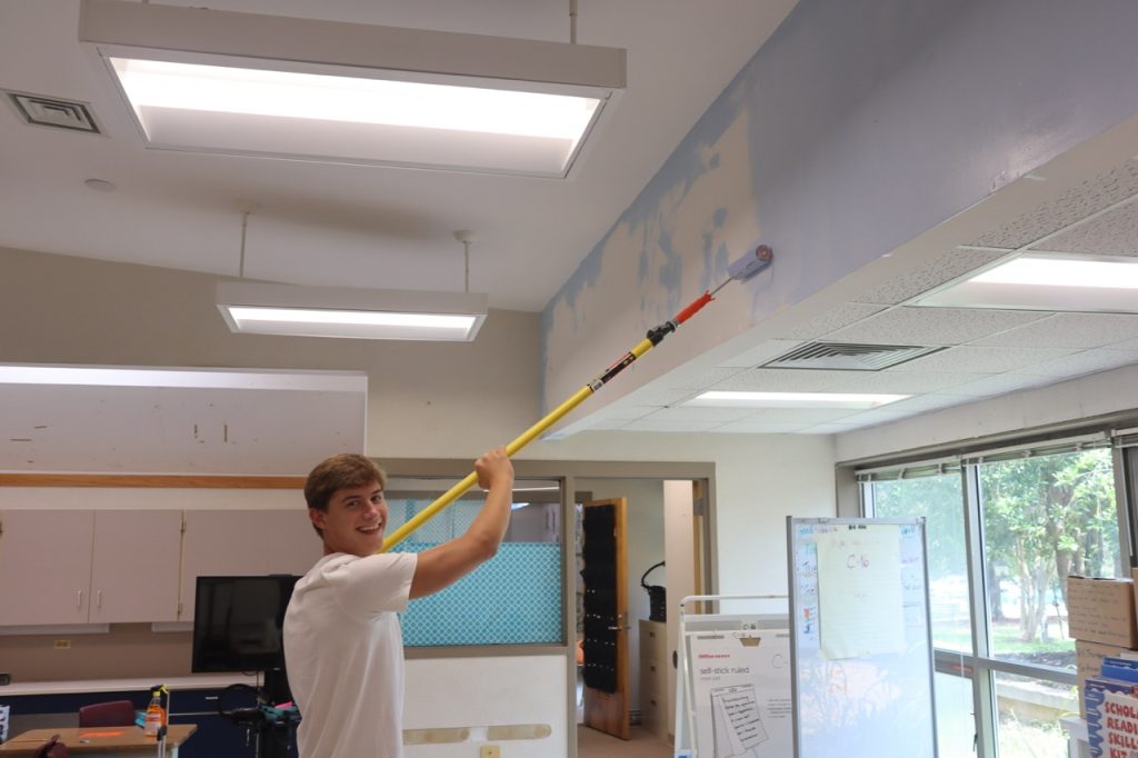 Student smiles as they use long paint roller to paint white wall light blue